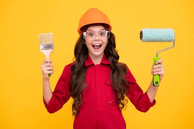 Child builder in hard hat helmet. Teenage girl painter with painting brush tool or paint roller. Teenager worker isolated on yellow background. Kids renovation concept. Excited teenager girl