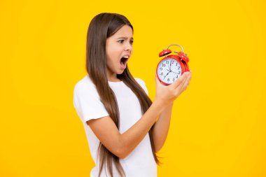 Teen girl holding clock over yellow background. Early morning, daily schedule. Child checking time. do not be late. Punctual kid with alarm. Deadline time. Angry face, upset emotions of teenager girl