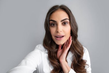 Portrait of cheerful smiling woman having video-call, shooting selfie. Young girl in white shirt with dark long hair isolated on gray background taking selfie photo, posing, smiling playful