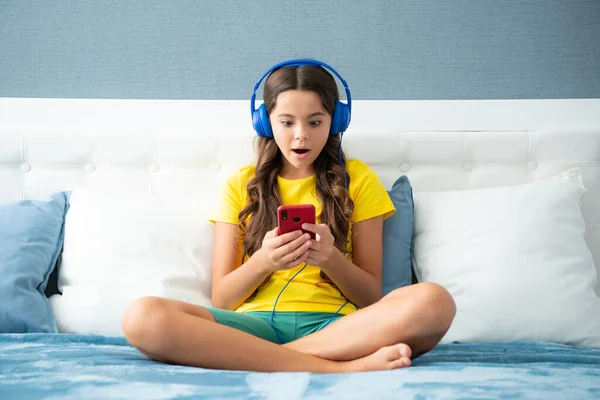 Teenager child girl wearing headphones watching videos on smart phone sitting on bed in her room. Surprised face, surprise emotions of teenager girl