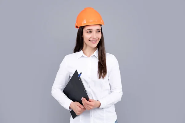 Young beautiful architect woman in hard hat helmet hold clipboard over gray background. Female builder in helmet for advertising sign.