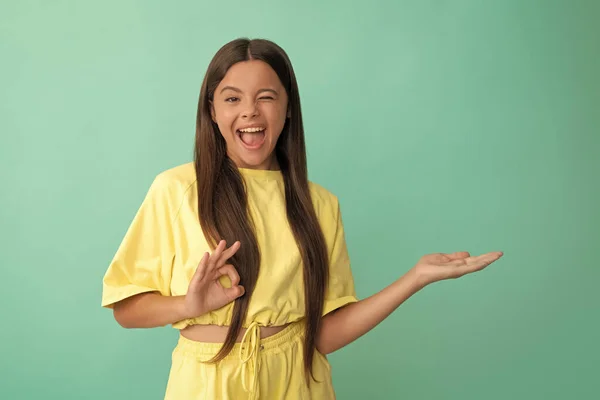 express positive emotions. ok. happy winking kid presenting product with copy space. beauty and fashion. female fashion model. pretty look of young girl. face portrait of child on blue background.