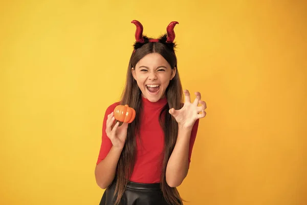 evil child in imp horns. happy halloween. devil kid with pumpkin. trick or treat. teen girl show claws on yellow background. celebrate autumn party holiday. childhood fun.