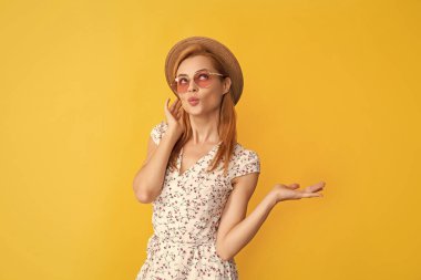 Sarı arka planda, hasır şapkalı ve güneş gözlüklü gülümseyen genç kadın..