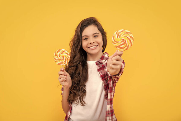 счастливый ребенок с длинными вьющимися волосами в клетчатой рубашке шоу леденец карамель конфеты на желтом фоне, стоматологическая помощь.