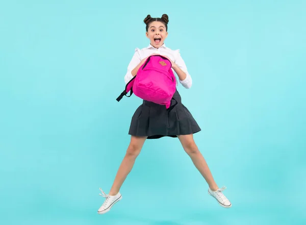 Amazed teenager. Back to school. Teenager schoolgirl in school uniform with backpack. Jump and run. School children on blue isolated studio background. Excited teen girl