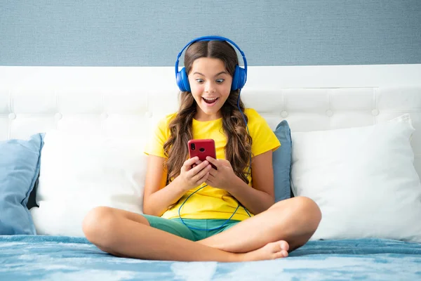 Surprised face, surprise emotions of teenager girl. Teenager child girl wearing headphones watching videos listening music on smart phone sitting on bed in her room