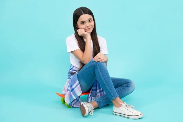Fashion girl wearing trendy t-shirt and jeans sit on skateboard, posing on blue background. Fashion teenager, casual look. Happy teenager, positive and smiling emotions of teen girl