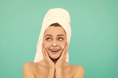 Şaşırmış genç bayan mavi arka planda yüz kremi sürüyor..