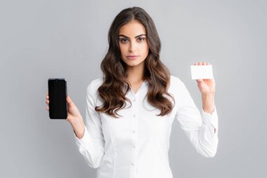 Amazed young brunette woman in shirt using mobile cell smart phone hold credit bank card isolated on gray background studio portrait
