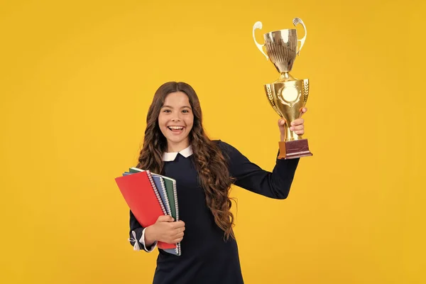 Excited schoolgirl in school uniform celebrating victory with trophy. Teen holding winning award prize on yellow background. Child hold book with trophy or winning cup. Education graduation, victory