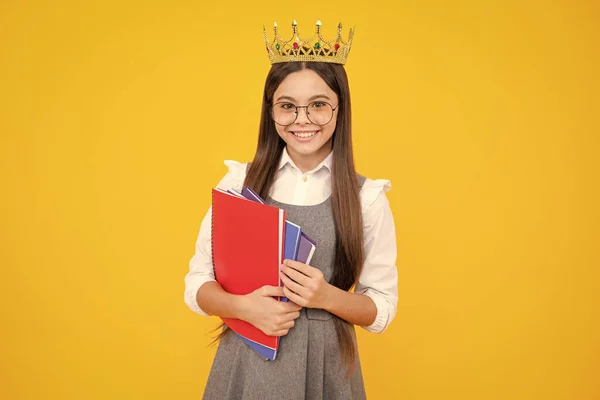 Schoolgirl in school uniform and crown celebrating victory on yellow background. School child hold books. Education graduation, victory and success