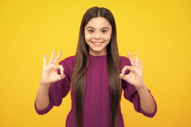 Güzel bir kız çocuğu sarı arka planda tamam işareti yapıyor. Gülümseyen çocuğun portresi kendine güvenen eller sembolü gösteriyor. Genç kız iyi niyet gösterisi yapıyor.
