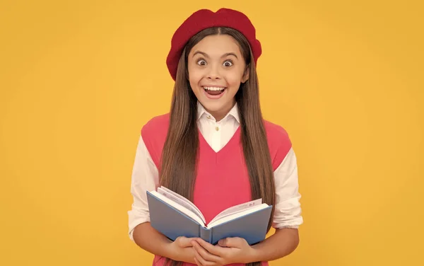 surprised teen school girl in french beret reading book on yellow background, happiness.