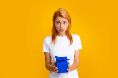 Surprised woman holding gift. Beautiful young woman in shirt hold present box with gift ribbon bow isolated on bright yellow background, studio portrait. Birthday concept