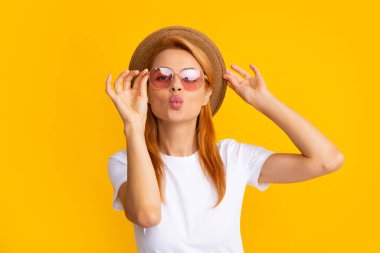 Young redhead woman in straw hat, surprised expression, isolated on yellow background. Summer lifestyle studio portrait. Redhead girl sends an air kiss