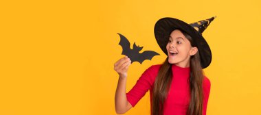 positive teen girl wearing witch hat holding bat on yellow background, childhood. Halloween kid girl portrait, horizontal poster. Banner header with copy space