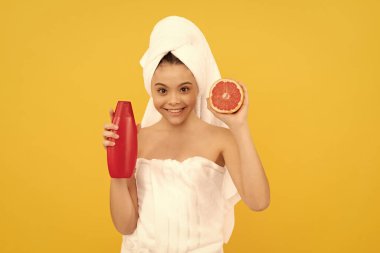 Havlu içinde, sarı arka planda greyfurt şampuanı şişesi olan mutlu bir çocuk..