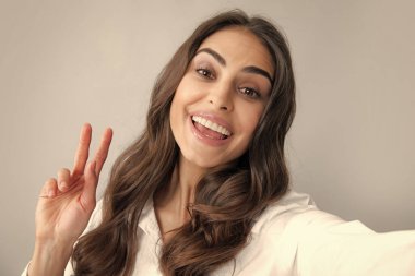Genç ve güzel bir kadının selfie çekerken yakın çekimi. İzole edilmiş gri arka plan. Barış işaretiyle poz veren güzel kız