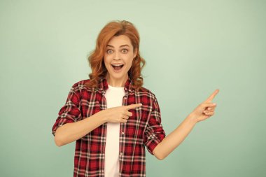 Şaşkın kızıl saçlı kadın mavi arka planda kareli gömlek parmağıyla işaret ediyor..