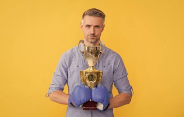 Boks eldivenli olgun bir adam sarı arka planda şampiyonluk kupasını tutuyor, başarılı..