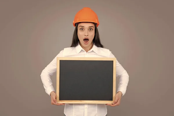 Surprised amazed wow woman construction manager. Woman builder isolated portrait with protect helmet and sign board. Gray background