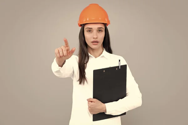 Architect woman in helmet pointing on copy space. Young woman in helmet hold clipboard on gray background. Female worker in hardhat helmet pointing to you