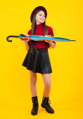 Fashion autumn kids weather, full length. Child teenager girl with umbrella in autumn skirt, sweater, boots and hat isolated on yellow studio background. Autumn trendy clothes