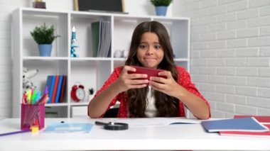 School child play smartphone games making winning gesture pretending doing homework at desk, mobile gaming.