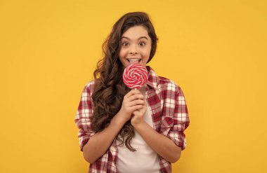 Kareli tişörtlü, uzun kıvırcık saçlı mutlu çocuk sarı arka planda lolipop karamel şekeri yalıyor..