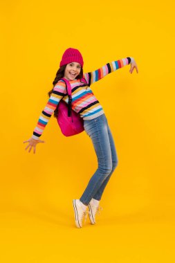 School children in winter hat and sweater with school bag on isolated yellow studio background. Children learning and education. Happy teenager, positive and smiling emotions of teen schoolgirl