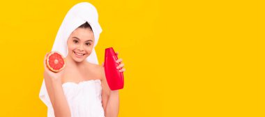 smiling kid in towel with grapefruit shampoo bottle on yellow background. Cosmetics and skin care for teenager child, poster design. Beauty kid girl banner with copy space