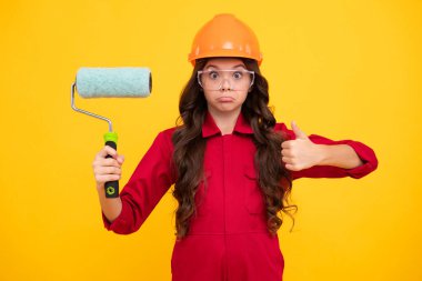 Funny face. Child builder in hard hat helmet. Teenage girl painter with painting brush tool or paint roller. Teenager worker isolated on yellow background. Kids renovation concept