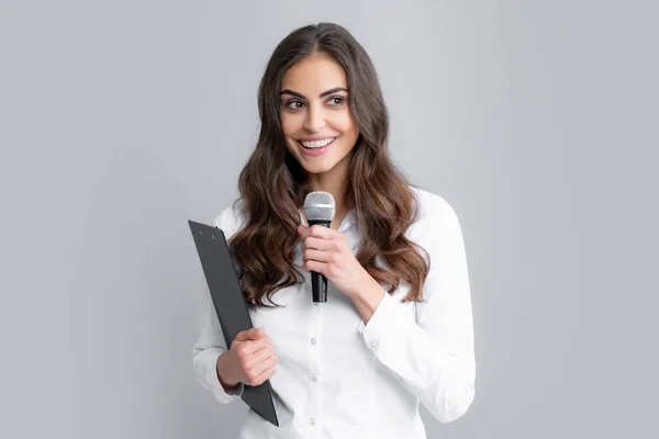 Portrait of young woman journalist in casual shirt holding microphone, asking questions, discussing problems, interviewing. Isolated on gray background