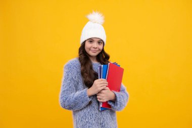 Sarı arka planda, kalın süveter ve şapka tutan kitaplar ve sırt çantası olan genç bir çocuk. Mutlu okul kızı, pozitif ve gülümseyen duygular