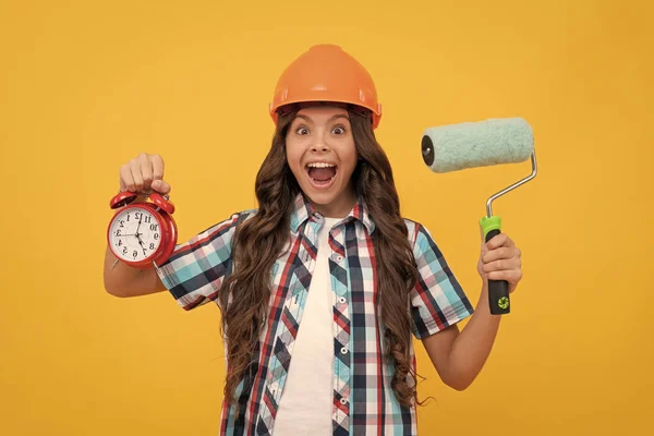 stock image amazed teen girl with curly hair in helmet hold paint roller and alarm clock, time to upgrade.