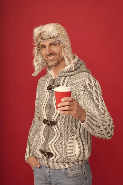 happy man in sweater and earflap drinking coffee from cup on red background, morning.