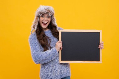 Genç kız, sıcak bir şapka içinde kara tahta tutarak sarı arka planda fotokopi alanı tutuyor. Heyecanlı bir yüz, neşeli genç bir kız.