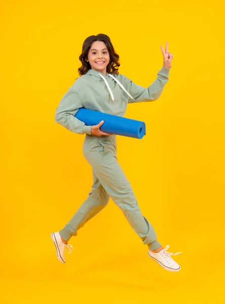 Run and jump. Full length of a fitness teen girl in sportswear hold yoga mat posing over yellow background. Fitness model child wearing sport clothes. Girl in the sport concept