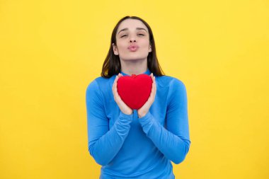 Aşkı hisseden bir kız. Mutlu kadın kırmızı kalbi tutar. İzole edilmiş sarı arka planda poz veren güzel bir kadın model. Neşeli, pozitif, tatlı, yumuşak kalpli bir kız.