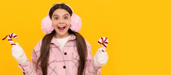 在新的一年里 戴着耳罩和手套的惊讶孩子们拿着黄色背景的棒棒糖 圣诞小女孩的横幅 带复制空间的工作室小孩冬季肖像 — 图库照片