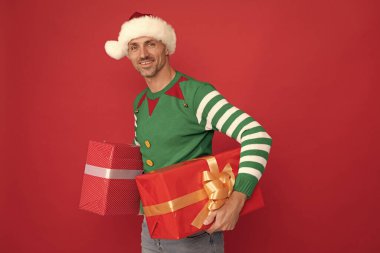 Elf kostümlü ve Noel Baba şapkalı mutlu bir adam. Kırmızı arka planda hediye kutusu olan xmas adamı. Mutlu yıllar. Mutlu noel hediyeleri.