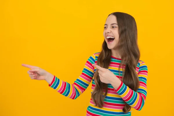 Positive teenager child points aside with cheerful expression, shows amazing at blank empty space. Excited face, cheerful emotions of teenager girl