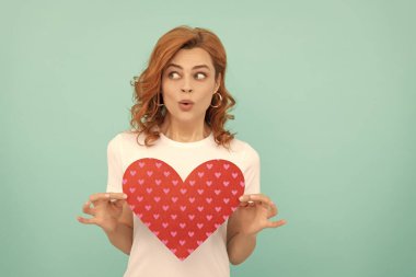 Şaşırmış kızıl saçlı kız mavi arka planda kırmızı kalbi tutuyor..