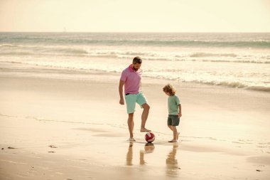 Spor aktivitesi. Baba ve oğul plajda futbol oynuyorlar. Baba ve çocuk güzel bir yaz gününde. Hafta sonu aile günü. Baba ve çocuk dışarıda eğleniyorlar. Çocukluk ve ebeveynlik. Aile tatilleri.
