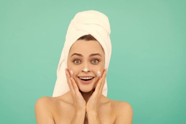 Şaşırmış genç bir kadın mavi arka plana yüz kremi sürmüş..