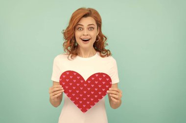 Şaşırmış kızıl saçlı kadın mavi arka planda kırmızı kalbi tutuyor..