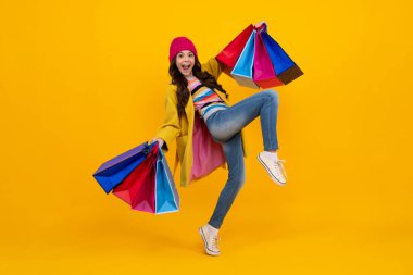 Sarı arka planda alışveriş torbaları olan güzel moda kızı. Alışveriş ve moda alışverişi. Koş ve atla. Alışveriş çantalı çocuk. Sonbahar alışverişi