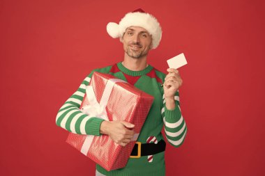 Alışveriş için bankamatik kartı tutan mutlu adam. Elf kostümlü ve Noel Baba şapkalı bir adam. Kırmızı arka planda hediye kutusu olan xmas adamı. Mutlu yıllar. Mutlu noel hediyeleri.