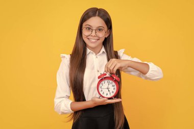 Okul üniformalı neşeli bir genç kız ve alarm saati takılı gözlükler sarı arka planda, okul saatinde.